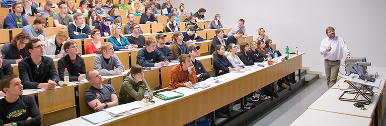 Ein Vortragender in einem Hörsaal erklärt Studierenden Inhalte